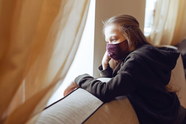 Concept of coronavirus quarantine. Child wearing protective face mask during flu virus, looking out of window. COVID-19 - self isolation. Teen girl forced to stay at home. Prevention epidemic.