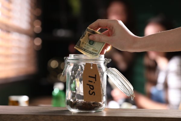 Woman,Putting,Tips,Into,Glass,Jar,On,Wooden,Table,Indoors,